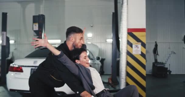 Przed kamerą dobrze wyglądający facet bawiący się z klientką na toczącym się krześle w nowoczesnym garażu samochodowym uśmiechają się — Wideo stockowe