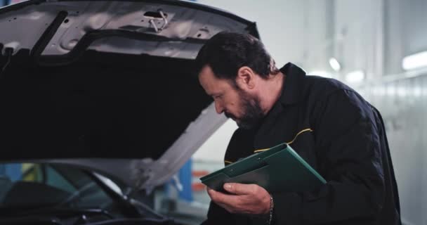 Smiling and happy mechanic man in a uniform while reading the documents from the map looking straight to the camera and feeling in a great mood in a auto service center — Stock Video
