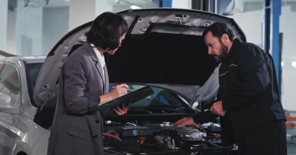 En un servicio moderno auto viejo mecánico chico con una señora gerente de oficina en un traje tienen una conversación que miran por encima del coche para encontrar el problema y a las notas en el mapa — Vídeo de stock