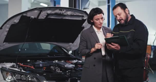 Ve velké moderní garážový mechanik muž v uniformě s mapou mají přátelský rozhovor s jeho zákazníkem podnikání žena se podívat na dokumenty a podepsat něco — Stock video