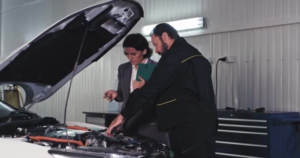 Attraktiver älterer Mechaniker in Uniform plaudert er mit seinem Büroleiter und analysiert das Problem vom Auto bis zur Reparatur in einem modernen Autoservice — Stockvideo