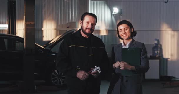 Gut aussehende Frau im Anzug vor der Kamera und reifer Mechaniker Mann, der gerade aussieht und lächelnde Dame mit einer Landkarte in einem Auto-Service-Center — Stockvideo