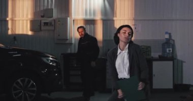 Harika bir ruh hali içinde mutlu bir kadın takım elbiseli ve otomobil servis merkezinde bir haritayla kamera önünde heyecanlı bir şekilde dans ediyor yetişkin bir tamirci de dans etmeye başlıyor.