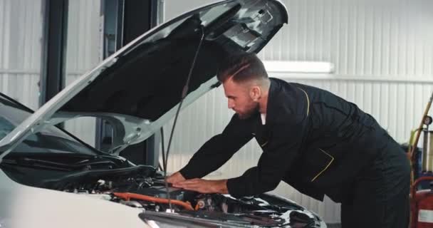 Potící se mechanik v garáži pracuje velmi soustředěný nad autem, opravuje problém, nosí černou speciální uniformu — Stock video