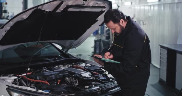 Mechanische man in uniform schrijven het probleem van de auto in een kaart in het moderne service center — Stockvideo