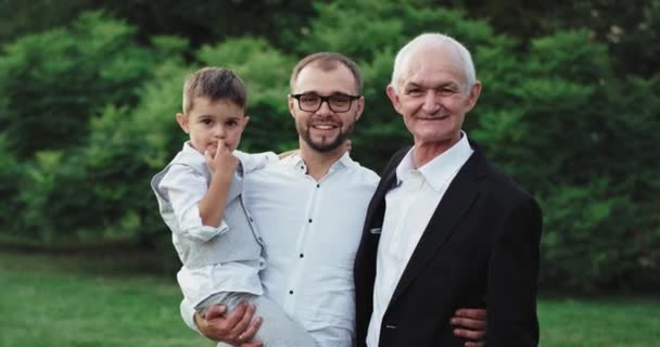 Посреди зеленого парка старик со своим сыном и внуком смотрит прямо в камеру семейный портрет улыбается мило — стоковое видео