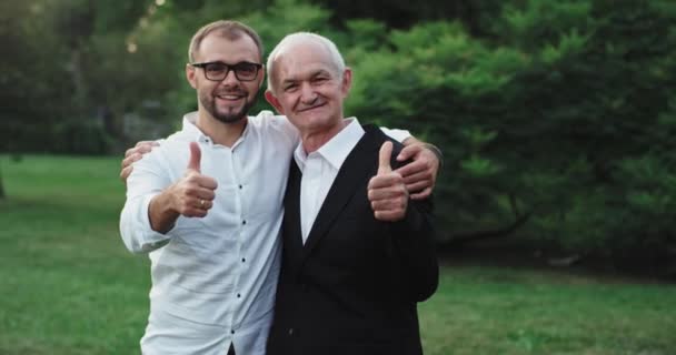 Two men old and young wearing casual clothes looking to the camera showing big like in the middle of a green park — Stock Video