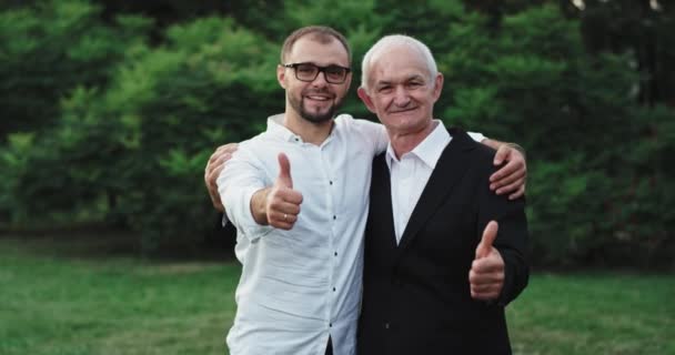 Attractive guy and his father in front of the camera showing a big like they stand up in the middle of the park — Stock Video