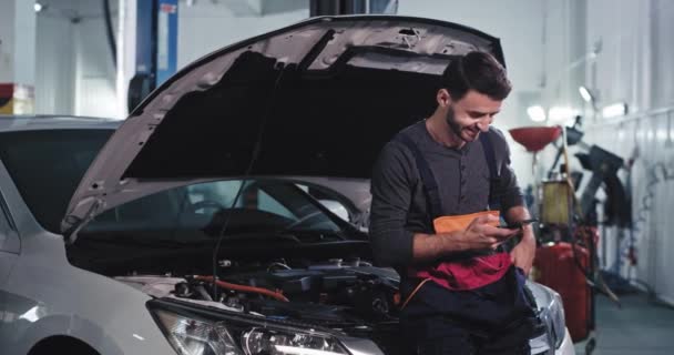 Usmívající se mechanik chlap v uniformě se podívat na jeho telefon při opravě auta v garáži