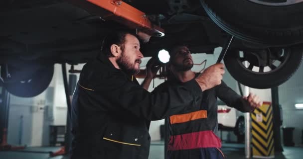 Jeden dospělý mechanik a jeho asistent v uniformě v garáži pod poškozeným autem se snaží najít problém, který je třeba opravit — Stock video