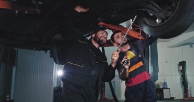 Otomobil hizmet merkezindeki kameraya yakın. Üniformalı iki tamirci el feneriyle hasarlı arabayı kontrol ediyor. İşe yoğunlaşmış.