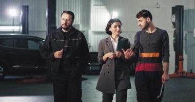 Bir oto hizmet merkezinde harika bir ekip üniformalı iki tamirci ve kameranın önünde yürüyen bir ofis çalışanı bir şeyi analiz ediyorlar.