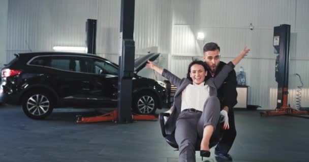 Happy business woman have a fun time while sitting on a rolling chair and a mechanic guy in a auto service pushing her they spend a great time after make a good deal — Stock Video