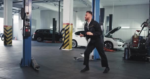 Dançando carismático cara mecânico na frente da câmera enquanto ele está no trabalho em um automóvel de serviço, consertando o carro — Vídeo de Stock