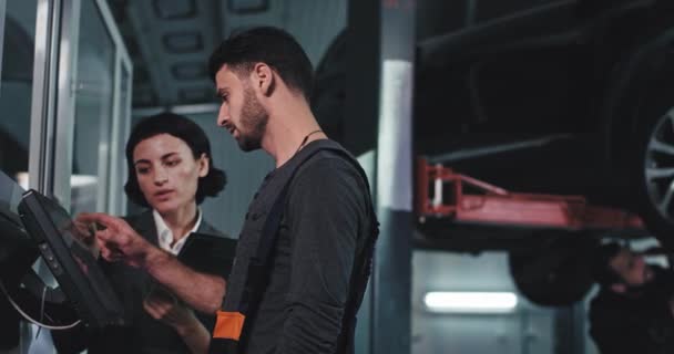 In a modern service center main mechanic guy in a uniform and the office manager woman analyzing something on the computer background other mature mechanic fixing the car — Stockvideo