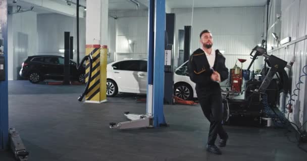 Homem mecânico em um uniforme dançando engraçado em um grande centro de auto serviço ele tem um ótimo humor no trabalho e ama seu local de trabalho — Vídeo de Stock