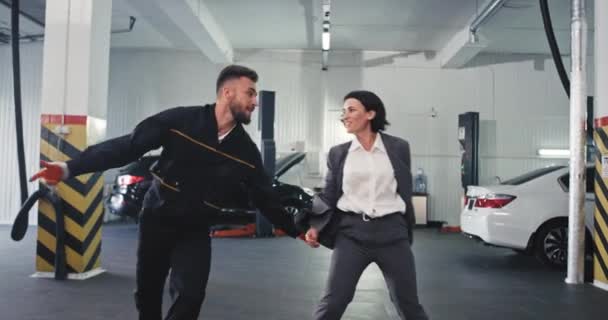 In a modern garage mechanic man with his customer lady dancing in front of the camera charismatic after they make a great deal — Stock Video