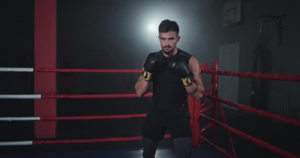 Bokser przed kamerą w ciemnej sali gimnastycznej w środku ringu bokserskiego — Wideo stockowe