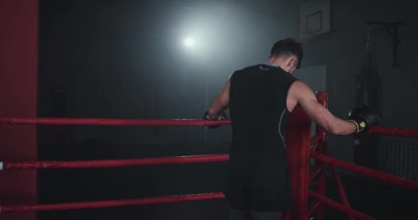 Na aula de ginástica escura no meio do ringue de boxe, o cara do corpo muscular atlético tem uma pausa depois de uma luta de boxe difícil. — Vídeo de Stock