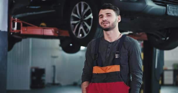 Beau mécanicien de voiture gars dans un uniforme profiter du temps au travail regardant droit à la caméra souriant grand et se sentant heureux — Video