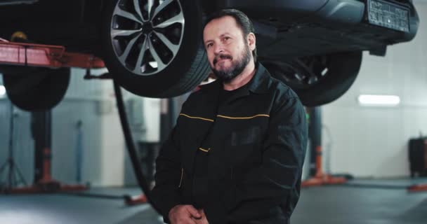Retrato delante de la cámara mecánico de coche maduro en un uniforme negro en un centro de servicio de coche moderno — Vídeos de Stock