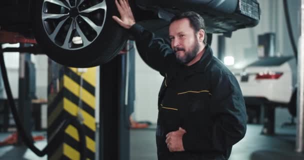 Gelukkig en glimlachend volwassen monteur in een auto service portret in de voorkant van de camera met een grote als terwijl naast een beschadigde auto — Stockvideo