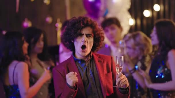 Jóképű göndör hajú fickó a kamera előtt pezsgőt iszik és élvezi a hangulatot a nagy partin nagyon izgatott és boldog. — Stock videók