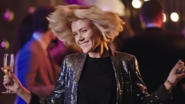 Счастливая и взволнованная блондинка, держащая бокал шампанского и танцующая взволнованная перед камерой на большой вечеринке, она качает головой, наслаждаясь моментом — стоковое видео
