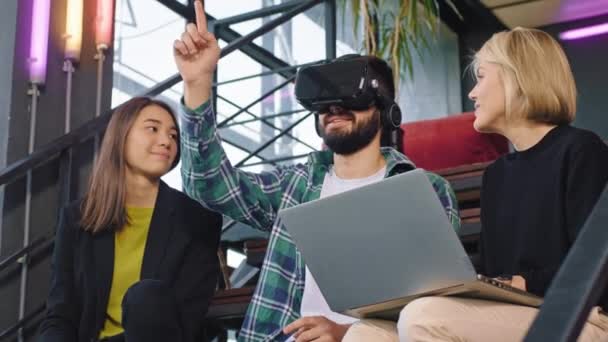 Chico guapo y dos señoras carismáticas usando gafas de realidad virtual para explorar el mundo mientras toman un café en su lugar de trabajo mientras están sentadas en las escaleras — Vídeo de stock