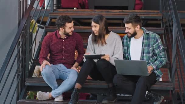 In einer modernen Bürotreppe drei attraktive Arbeiter zwei Jungs und eine Dame analysieren den Arbeitsplan, während sie auf der Treppe sitzen — Stockvideo