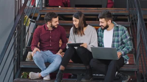 Na nowoczesnych schodach biurowych trzech atrakcyjnych pracowników dwóch facetów i jedna pani analizująca plan pracy siedząc na schodach — Wideo stockowe