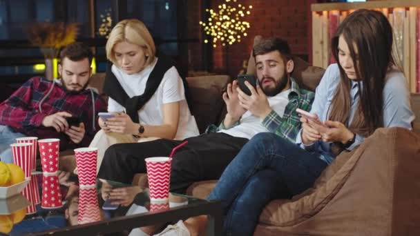 Geconcentreerde charismatische mooie jonge mensen thuis feest in een moderne stijlvolle woonkamer zittend op de bank en met behulp van smartphone besteden geweldige tijd — Stockvideo