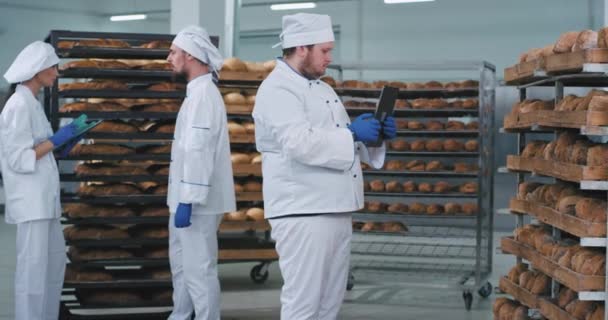 In a bakery industry main baker man taking photos from a new technology tablet of a baked fresh bread from shelves other two workers chatting on the background — ストック動画