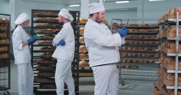 I en stor bageriindustri tar huvudbagaren foto från en stor platta med ett nybakat bröd från hyllorna medan andra två bagare chattar på bakgrunden. 4k — Stockvideo