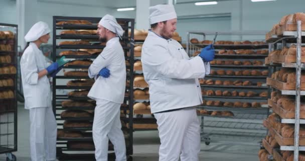 Ve velkém pekařském průmyslu hlavní pekař fotit z velké tablety čerstvě pečený chléb z regálů, zatímco další dva pekaři klábosí na pozadí. 4k