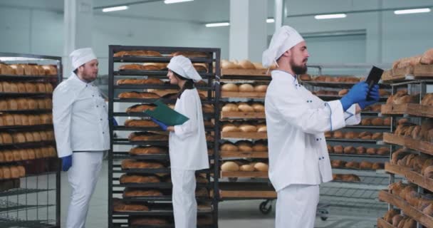 In een bakkerij industrie neemt de bakker enkele notities en foto 's met behulp van een tablet andere twee bakkers chatten op de achtergrond in een grote bakkerij sectie vol vers gebakken brood op planken — Stockvideo