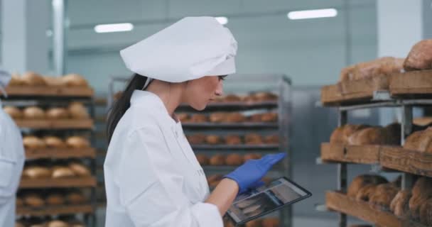 Good looking lady with a new big tablet take some photos of a fresh baked bread from the shelves in a big bakery factory in the background two other bakers chatting. 4k — ストック動画