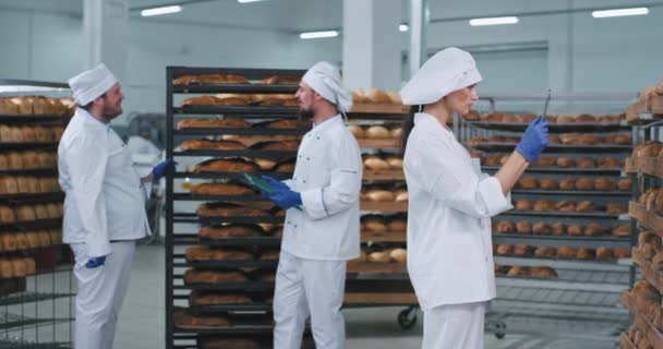 Велике відео хлібопекарської промисловості приймає три пекарі спілкуючись один з одним тон красива пані пекарня в білій формі фотографії свіжого запеченого хліба — стокове відео