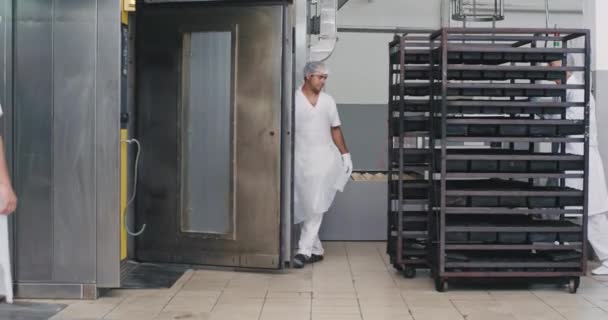 Stora bageriarbetare i vit uniform lasta hyllorna med råa bröd på industriugnen för att få bakning, livsmedelsindustrin tillverkning — Stockvideo