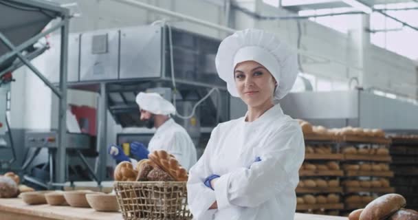 Женщина-пекарь мило улыбается перед камерой на фоне крупной пекарни и булочника, смешно двигающегося. 4k — стоковое видео