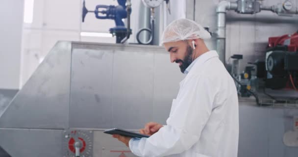 Good looking engineer in a big manufacturing industry holding a electronic tablet and start to checking the industrial machine , he wearing the wireless headphones — Stock Video