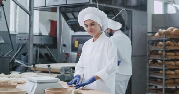 Симпатичная зрелая женщина с большой улыбкой пекаря в белой форме готовит тесто для выпечки хлеба в то время как другие работники проверить машину и переместить полки — стоковое видео