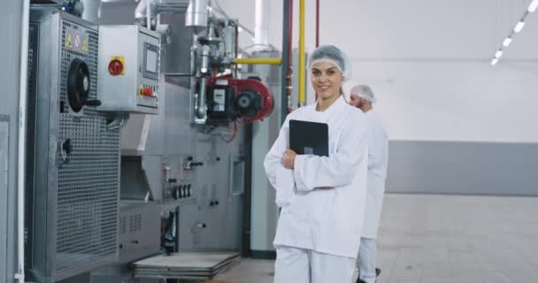 Snygg ingenjör kvinna med ett perfekt leende i en stor tillverkningsindustri håller en elektronisk tablett tittar rakt mot kameran och ler bakgrund annan ingenjör kontrollera — Stockvideo
