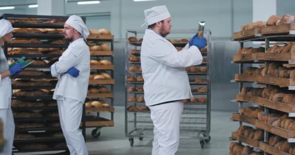 Hombre panadero principal utilizando una tableta de nueva tecnología para tomar fotos de pan recién horneado de estantes otros dos trabajadores hablando en el fondo — Vídeo de stock