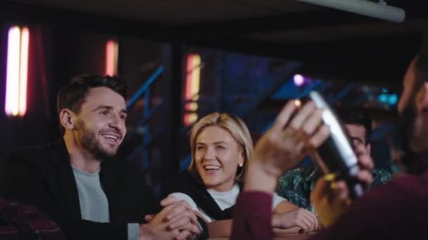 Sonriente grupo de amigos carismáticos en un bar moderno mientras se sientan en la mesa del bar y esperan sus cócteles mirando al camarero mientras prepara las bebidas . — Vídeos de Stock