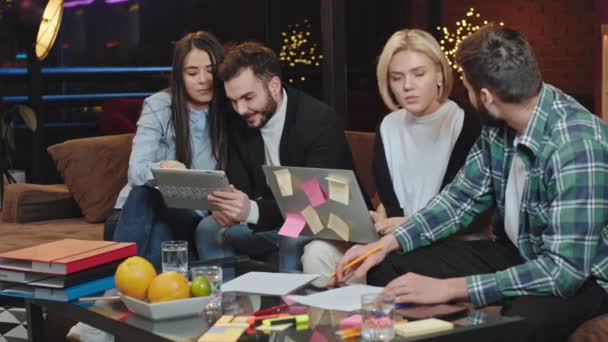 Munkavállalók csoportja egy irodai modern stúdióból, akik úgy dolgoznak együtt, mint egy csapat, miközben a kanapén ülnek jegyzetfüzettel és elektronikus táblagéppel — Stock videók