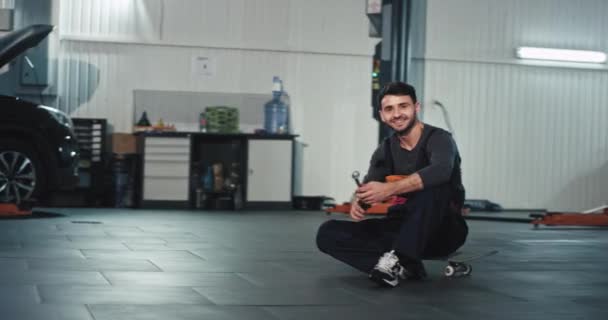 Carismático jovem mecânico em um uniforme sorrindo grande e olhando direto para a câmera e mostrando um grande como enquanto ele se senta no skate — Vídeo de Stock