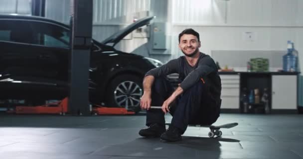 Mechanik chlap se usmívá a atraktivní v uniformě před kamerou pocit, šťastný, zatímco sedí na svém skateboardu — Stock video