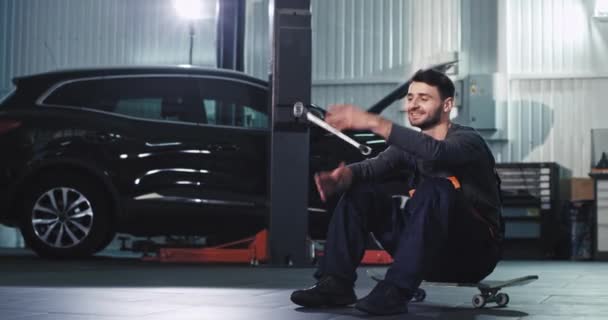 In front of the camera smiling and happy mechanic in a uniform excited sitting down on the skateboard and feeling great — Stock Video