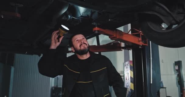 In un meccanico di buon umore che usa una torcia sotto l'auto danneggiata in garage cerca di trovare il problema. — Video Stock
